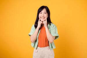 vrolijk Aziatisch vrouw Jaren 30 vervelend groen en oranje shirt. met vingers richten naar tanden, benadrukt tandheelkundig Gezondheid en stralend glimlach geïsoleerd Aan geel achtergrond. bevorderen mondeling zorg concept. foto