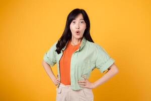 expressief en verrassend, een jong Aziatisch vrouw Jaren 30 vervelend een groen overhemd Aan een oranje overhemd tonen verrassing gezicht terwijl staand met akimbo armen. niet verwacht met levendig beeld Aan geel achtergrond. foto