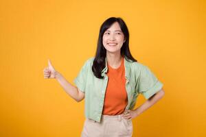 jong Aziatisch vrouw in haar jaren '30, aandoen een oranje overhemd en groen trui. haar duimen omhoog gebaar, geïsoleerd Aan een levendig geel achtergrond, betekent: een concept van positiviteit en overeenkomst. foto