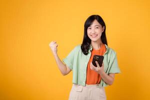 opgewonden jong Aziatisch vrouw in haar jaren '30, vervelend oranje overhemd en groen trui, gebruik makend van smartphone met vuist omhoog gebaar Aan geel studio achtergrond. Super goed nieuws van mobiel concept. foto