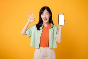 nieuw mobiel toepassing met een levendig jong Aziatisch vrouw in haar jaren '30, gekleed in oranje overhemd en groen trui, presentatie van smartphone scherm met een Oke hand- gebaar Aan geel studio achtergrond. foto