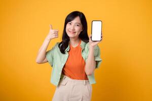 nieuw mobiel toepassing met een vrolijk jong Aziatisch vrouw in haar jaren '30, aandoen oranje overhemd en groen trui, weergeven smartphone scherm met een duimen omhoog gebaar Aan geel studio achtergrond. foto