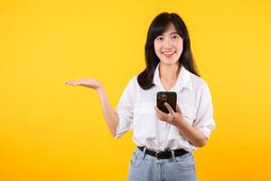 portret mooi jong Aziatisch vrouw gelukkig glimlach vervelend wit overhemd en denim planten tonen smartphone met richten hand- gebaar naar vrij ruimte geïsoleerd Aan geel achtergrond. app smartphone concept. foto