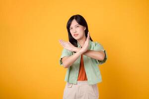 ontkenning met een jong Aziatisch vrouw in haar jaren '30, gekleed in een oranje overhemd en groen trui. haar kruis hand- gebaar, geïsoleerd Aan een levendig geel achtergrond, belichaamt de concept van weigering en negatie. foto