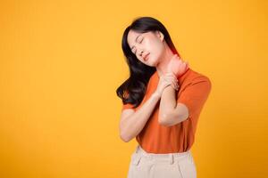 bezorgd Aziatisch vrouw in haar jaren '30, vervelend een oranje shirt, houdt haar pijnlijk nek Aan geel achtergrond. kantoor syndroom Gezondheid zorg concept. foto