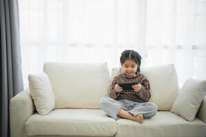 Aziatisch kind meisje op zoek gebruik makend van en tintje mobiel telefoon scherm Aan bankstel bank. baby glimlachen grappig tijd naar gebruik mobiel telefoon. te veel scherm tijd. schattig meisje aan het kijken videos terwijl TV, internet verslaving. foto