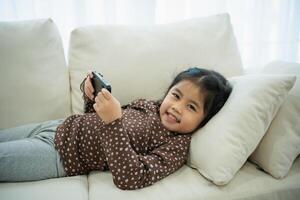 Aziatisch kind meisje op zoek gebruik makend van en tintje mobiel telefoon scherm Aan bankstel bank. baby glimlachen grappig tijd naar gebruik mobiel telefoon. te veel scherm tijd. schattig meisje aan het kijken videos terwijl TV, internet verslaving. foto