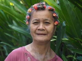 een ouderen Aziatisch vrouw met haar- rol, glimlachen en op zoek Bij de camera terwijl staand in een tuin foto