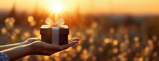 ai gegenereerd hand- Holding een geschenk doos in een zonsondergang backdrop net zo een gebaar van geven. een hand- strekt zich uit uit aanbieden een doos verpakt in papier en gebonden met een lintje. foto
