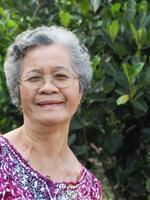 senior vrouw met kort wit haar- glimlachen en op zoek Bij camera terwijl staand in een tuin foto