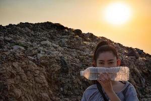 vrouwenhand die een doorzichtige plastic fles vasthoudt om het gezicht te bedekken op de grote vuilnisachtergrond van de berg foto