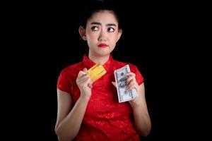 portret van een mooie jonge azië vrouw rode jurk traditionele cheongsam met creditcard en geld bankbiljet 100 usd op de zwarte achtergrond foto