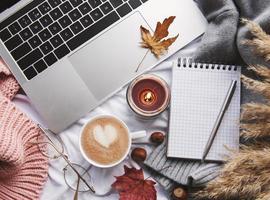 laptop, trui, gele herfstbladeren, kaars en koffie op wit bed.