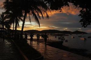 zonsondergang aan de waterkant. silhouetten van palmbomen, mensen die wandelen en genieten van het strandlandschap, boten verankerd aan de kust. foto