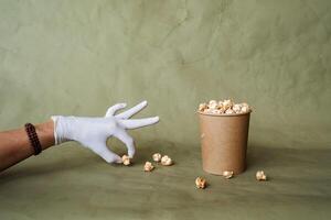 hand- in een wit handschoen bereikt voor popcorn, nemen voedsel in beschermend handschoenen, houden een stuk van voedsel met twee vingers, een vol doos van popcorn, veiligheid Bij de snel voedsel verdeling, minimaal concept foto