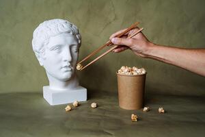 voeden een gips hoofd met popcorn van een emmer, kunst concept, creativiteit, snel voedsel en Chinese eetstokjes foto