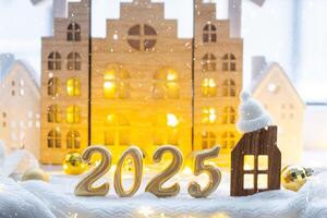 gouden figuren aantal 2025 en klein huis Aan achtergrond van knus ramen van een huis met warm licht met feestelijk decor van sterren, sneeuw en slingers. groet kaart, gelukkig nieuw jaar, knus huis foto