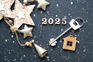 huis sleutel met sleutelhanger huisje Aan feestelijk zwart achtergrond met sterren, lichten van slingers. nieuw jaar 2025 houten brieven, groet kaart. aankoop, bouw, verhuizing, hypotheek, verzekering foto