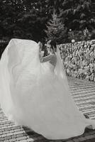 de bruid in een luxueus jurk en sluier wandelingen naar beneden de steen pad met haar schouders gedraaid. zwart en wit foto. prachtig jurk met lang mouwen, Open borstbeeld. zomer bruiloft foto