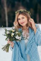 portret van een schattig bruid in een wit jurk met een boeket van bloemen in haar handen, verpakt in een blauw plaid, aan het wachten voor de bruidegom tegen de achtergrond van een winter landschap van met sneeuw bedekt bergen. foto