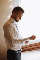 de bruidegom toetsen zijn overhemd door de venster. Mens in een shirt. mannetje portret. fotoshoot Bij de venster. bruidegom verzameling. bruiloft dag. knap Mens bruidegom knopen zijn overhemd in een hotel kamer. foto