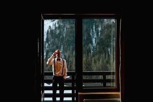 voorkant visie van een Mens in bretels en een boog stropdas staand Aan een balkon met uitzicht met sneeuw bedekt bergen. rust uit in de bergen. berg lucht. bruiloft in winter foto