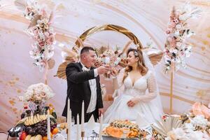 mooi bruid en bruidegom vieren hun bruiloft Bij een feest. pasgetrouwden maken een geroosterd brood naar een gelukkig huwelijk, staand Bij een dining tafel versierd met bloemen en droog bloemen foto
