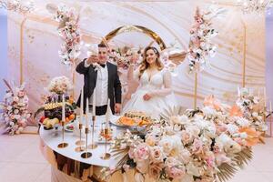 mooi bruid en bruidegom vieren hun bruiloft Bij een feest. pasgetrouwden maken een geroosterd brood naar een gelukkig huwelijk, staand Bij een dining tafel versierd met bloemen en droog bloemen foto