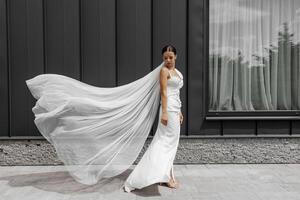 mooi jong bruid meisje vervelend wit jurk met lang sluier en poseren in de buurt grijs muur in modern stad buitenshuis onder zonlicht. foto