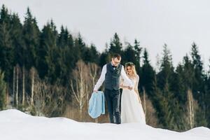 een mooi paar is staand in een winter pijnboom Woud, omringd door een veel van sneeuw. de bruid in een wit bruiloft jurk, de bruidegom in een wit overhemd en vest. winter bruiloft concept. foto