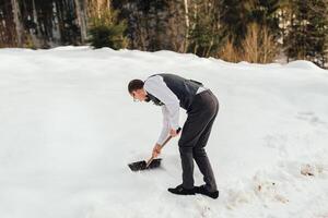 een knap Mens schoppen sneeuw in de bergen, bereidt zich voor rodelen gronden foto