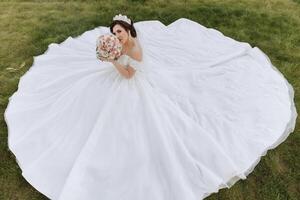 brunette bruid in off-the-schouder kant wit jurk en tiara poseren met een boeket van wit en roze bloemen terwijl zittend Aan groen gras. mooi haar- en verzinnen. voorjaar bruiloft foto