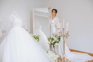 een gelukkig bruid is voorbereidingen treffen voor haar luxueus bruiloft in een hotel kamer, met een bruiloft jurk Aan een mannequin in de buurt. portret van een vrouw met modieus haar, bedenken en een glimlach in een dressing gewaad. foto