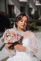 brunette bruid in off-the-schouder kant wit jurk en tiara poseren met een boeket van wit en roze bloemen. mooi haar- en verzinnen. voorjaar bruiloft foto