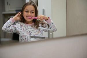 reflectie in de badkamer spiegel van een schattig kind meisje poetsen tanden, gekleed in pyjama. tandheelkundig hygiëne en mondeling zorg foto