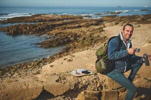 gelukkig Mens avonturier zit door atlantic strand, glimlacht op zoek Bij camera, houdt een thermosfles, geniet zijn koffie breken buitenshuis foto