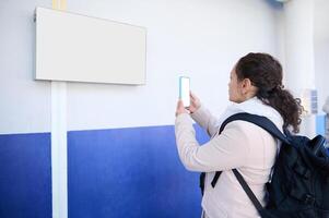 vrouw forens met rugzak, fotograferen vertrek bord met trein schema's, staand Bij spoorweg station platform foto