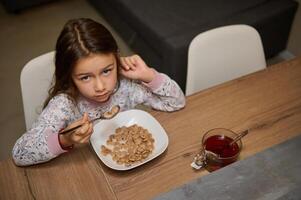 een weinig kind meisje in elegant pyjama, zittend Bij keuken tafel en aan het eten haar gezond ontbijt in de ochtend- foto