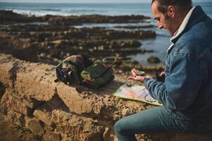 gelukkig mannetje toerist avonturier met kompas en verrekijker, zittend Aan de rots door zee en lezing navigatie kaart foto
