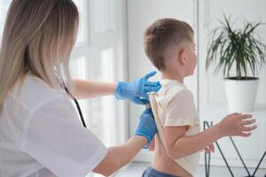 de dokter luistert naar de ademen van een weinig jongen door een stethoscoop. kinderarts met stethoscoop luistert naar de longen van een kind met bronchitis en hoesten foto