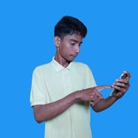 jong Aziatisch Mens verrast op zoek Bij slim telefoon geïsoleerd blauw achtergrond. vervelend een geel t-shirt foto