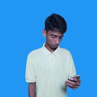 jong Aziatisch Mens verrast op zoek Bij slim telefoon geïsoleerd blauw achtergrond. vervelend een geel t-shirt foto