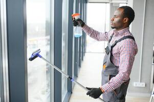 arbeider van schoonmaak organisatie voorzichtig en voorzichtig wrijft groot venster van de kantoor ruimte. een echt Afro-Amerikaans in blauw overall doekjes de dubbel glas venster in de kantoor foto