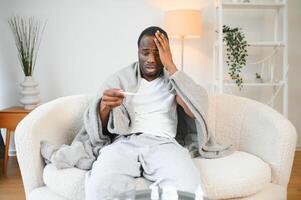 koorts. portret van ziek Afrikaanse Amerikaans Mens met thermometer lijden van griep symptomen zittend verpakt in deken in bed Bij huis foto