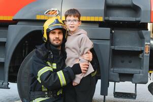 een brandweerman nemen een weinig kind jongen naar opslaan hem. brand motor auto Aan achtergrond foto