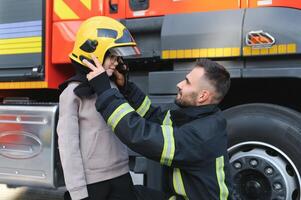 een brandweerman shows zijn werk naar zijn jong zoon. een jongen in een brandweerman helm foto