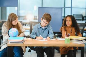 gefocust multiraciaal studenten kinderen schrijven naar beneden gegevens in notitieboekje terwijl zittend Bij tafel foto