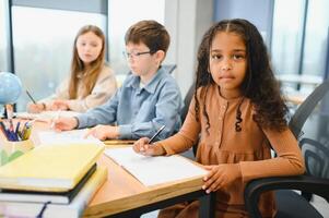 Afrikaanse Amerikaans school- meisje schrijven aan het leren zittend Bij bureau in klas binnenshuis foto