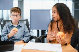 multiraciaal schoolkinderen hebben lunch Bij de bureau gedurende een breken in school- foto