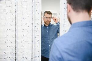 in optiek winkel. portret van mannetje cliënt Holding en vervelend verschillend bril, kiezen en proberen Aan nieuw bril Bij optisch op te slaan. Mens plukken kader voor visie correctie, detailopname. foto